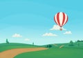 Striped hot air balloon flying over rural landscape with green meadows, trees, bushes, dirt road and forest with clouds and blue