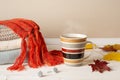 Striped mug of hot steaming coffee or tea, stack of warm knitted sweaters and red fringed scarf and few yellow fall leaves over