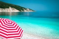 Striped red sun beach umbrella on a pebble beach with turquoise blue sea, white rocks and sky in background Royalty Free Stock Photo