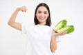 Strong asian woman shows fresh vegetables, lettuce and her muscles, flexing biceps with smiling face, white background