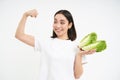Strong asian woman shows fresh vegetables, lettuce and her muscles, flexing biceps with smiling face, white background