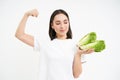 Strong asian woman shows fresh vegetables, lettuce and her muscles, flexing biceps with smiling face, white background