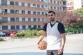 Strong and attractive basketball player looks at camera smiling while posing with his ball on an urban court