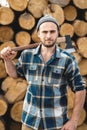 Strong bearded woodcutter holds axe on his shoulder on warehouse of logs