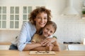 Affectionate young woman parent cuddle girl child by kitchen table