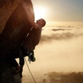 Brave male climber blinded by sunlight on high natural rock wall over gray clouds. Climbing with rope and safety harness