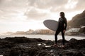 Strong brunette boy standing in the black swimsuit with a white surf in his hands on the shore Royalty Free Stock Photo