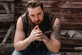 Strong brutal man with a beard and tattoos on his hands dressed in leather vest and jeans sits on a wooden wall