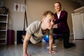 Strong male patient doing pushups with black rubber band while female practitioner supports, kneeling in exercise studio