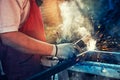 Strong man is a welder in T-shirt in a construction gloves, a metal product is welded with a welding machine in the garage