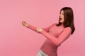 Strong purposeful woman with brown hair in pink sweater making great efforts to achieve success, pretending to pull rope