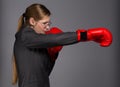 Strong and resolute young woman in dark grey business suit, glasses and red boxing gloves stands in fighting pose with punch