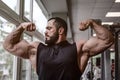 Strong young athlete man with beard wearing black tank top showing big double biceps muscle in sport gym with window Royalty Free Stock Photo