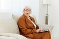 struck with surprise and shock, an emotional woman with age-gray hair sits on the couch with a laptop while working from