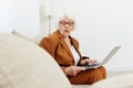 struck with surprise and shock, an emotional woman with age-gray hair sits on the couch with a laptop while working from