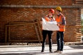 Structural engineer and architect dressed in orange work vests and helmets discuss documentation on a brick wall