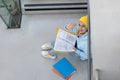 A student in a blue sweatshirt and yellow hat sits with folders between the stairs and greets a friend