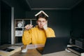 Student with a book on his head sleeps sitting at a table at home in the bedroom with books, notebook, laptop and pen in hand. Royalty Free Stock Photo