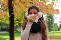 Student girl with earmuffs and scarf sneezing and blowing nose into tissue in city park