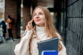 student girl talks on a mobile phone holds folders, books, notebooks, smiles, looks up a on the background of a modern Royalty Free Stock Photo