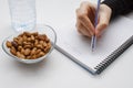 Student hand holding pen and writing notes in the notebook and glass bowl of hazelnuts and water bottle Royalty Free Stock Photo