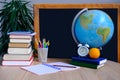 Student home office table with white alarm clock, books, orange, colored notebooks, pencils in glass, chalk board, globe, white Royalty Free Stock Photo