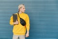 Student with a laptop in his hands and a fanny pack around the neck standing against the background of a blue wall and looking