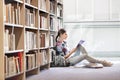 Student reading book while sitting against bookshelf at library Royalty Free Stock Photo