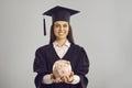 Happy student in academic cap and gown holding piggy bank with money saved for education Royalty Free Stock Photo