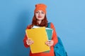 Student showing paper folder to camera, looks happy and satisfied, has rucksack on back, wearing orange jumper and cap, student Royalty Free Stock Photo