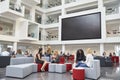 Students sit talking under AV screen in atrium at university