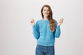 Studio portarit of funny and caucasian adult female in trendy sweater standing with lifted hands over gray