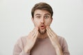 Studio portrait of amazed young man with bristle touching his face with surprised expression, standing against gray Royalty Free Stock Photo