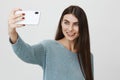 Studio portrait of attractive happy girl taking selfie with new smartphone, smiling brightly while standing over gray Royalty Free Stock Photo