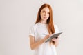 Studio portrait of attractive redhead young woman writing in diarylooking at camera, standing on white isolated Royalty Free Stock Photo