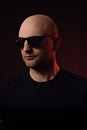 Studio portrait of a bald unshaven man on dark background in dark t shirt and glasses. Colored rim light