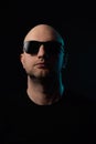 Studio portrait of a bald unshaven man on dark background in dark t shirt and glasses. Colored rim light