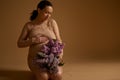 Studio portrait of gorgeous pregnant woman putting her hand on belly, holding bunch of purple lilacs, isolated on beige Royalty Free Stock Photo