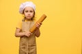 Adorable little kid girl in chef's hat and apron, stading with a rolling pin, isolated over yellow studio background Royalty Free Stock Photo