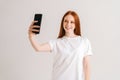 Studio portrait of cheerful redhead young woman taking selfie and smiling using smartphone standing on white isolated Royalty Free Stock Photo