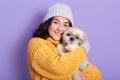 Studio portrait of cute young woman wearing cap and yellow sweater, looks smiling at camera and hugging her puppy. Girl playing Royalty Free Stock Photo