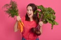 Studio portrait of emotional cheerful young vegetarian holding beet and carrots, opening mouth widely, looking at vegetables,