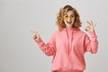 Studio portrait of emotive happy feminine woman with curly hair, wearing casual hoodie and sunglasses showing ok sign