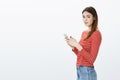 Studio portrait of gorgeous feminine european woman with brown hair, standing in profile, holding smartphone and looking