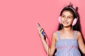 Studio portrait of happy little child girl, holding smartphone in hands, using headphones. Background of pastel pink color.