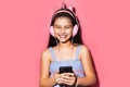 Studio portrait of happy little child girl, holding smartphone in hands, using headphones. Background of pastel pink color.