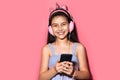 Studio portrait of happy little child girl, holding smartphone in hands, using headphones. Background of pastel pink color.