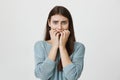 Studio portrait of horrified and shocked girl holding hands near mouth and looking afraid at camera, standing over gray Royalty Free Stock Photo