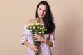 Studio portrait of miling Caucasian woman wearing pink dress, holding bouquet of flowers over beige background,