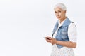 Studio portrait modern granny with combed grey hair standing in profile over white background, hold smartphone, reading